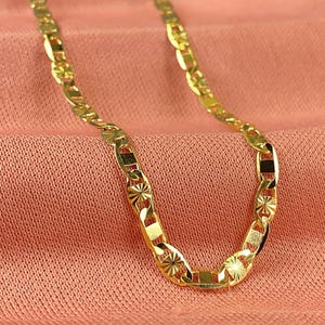 May include: A gold-colored chain necklace with a unique link design, resting on a peach-colored fabric. The chain features starburst and rectangular details. The image also includes a logo with the image of the Virgin Mary.