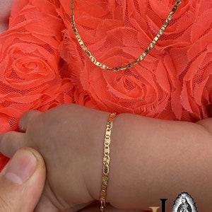 Puede incluir: Un conjunto de collar y pulsera de color dorado. El collar es una cadena delicada con un patr&oacute;n repetitivo, y la pulsera tiene un dise&ntilde;o similar. Las joyas se muestran en una persona que lleva un vestido color coral.