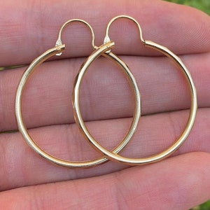 May include: A pair of gold hoop earrings with a hammered texture. The earrings are shown on a hand against a green background.