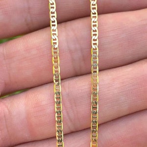May include: A gold-colored mariner chain necklace. The chain is made of small, rectangular links. The necklace is held in a person's hand. The background is blurred green.
