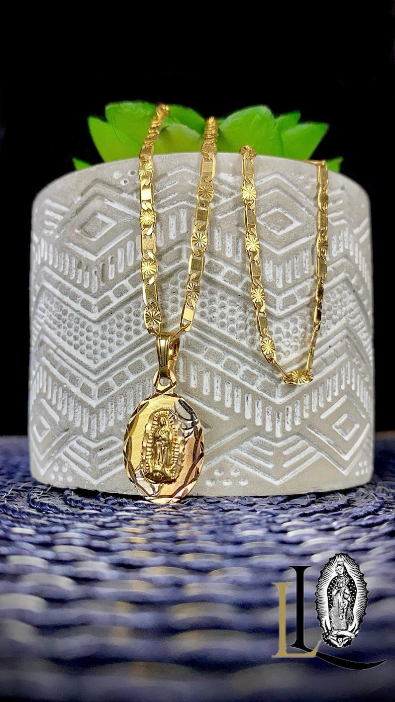 May include: A gold chain necklace with a pendant featuring an image of the Virgin Mary. The chain is a box chain with a diamond-cut design. The pendant is oval-shaped and has a detailed image of the Virgin Mary.