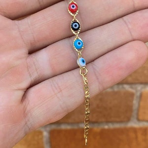 May include: A gold chain bracelet with five evil eye charms. The charms are blue, red, black, turquoise, and white.