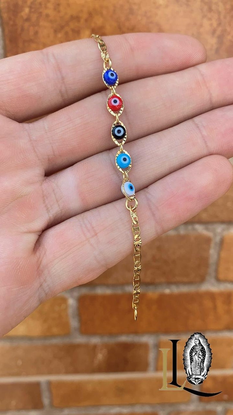 May include: A gold chain bracelet with five evil eye charms. The charms are blue, red, black, turquoise, and white.
