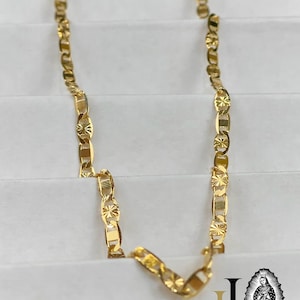 May include: A gold-colored chain necklace with a repeating pattern of rectangular and star-shaped links. The necklace is displayed on a white surface. The image also includes a logo with the image of a religious figure.