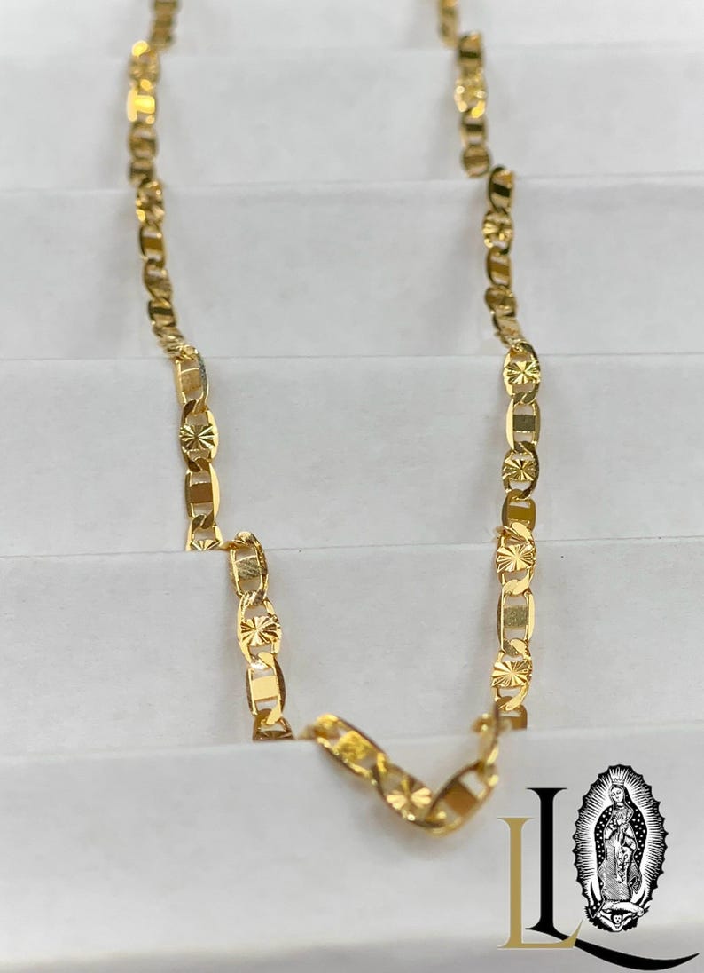 May include: A gold-colored chain necklace with a repeating pattern of rectangular and star-shaped links. The necklace is displayed on a white surface. The image also includes a logo with the image of a religious figure.