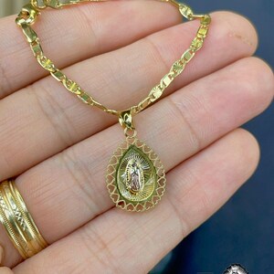 Puede incluir: Collar dorado con un colgante que presenta una figura religiosa. El colgante tiene forma de l&aacute;grima con un borde en forma de coraz&oacute;n. El collar se muestra en una mano, con un anillo de oro visible.