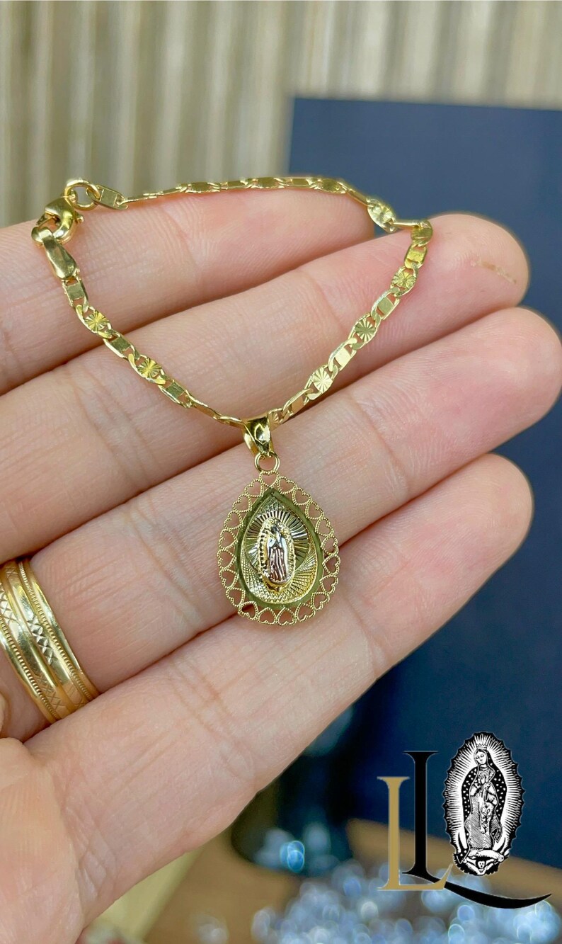 Puede incluir: Collar dorado con un colgante que presenta una figura religiosa. El colgante tiene forma de l&aacute;grima con un borde en forma de coraz&oacute;n. El collar se muestra en una mano, con un anillo de oro visible.