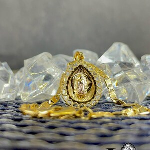 Puede incluir: Un collar con colgante dorado que representa a la Virgen Mar&iacute;a. El colgante tiene un dise&ntilde;o calado y ornamentado y cuelga de una cadena a juego. El collar se exhibe sobre una superficie azul texturizada con detalles transparentes en forma de cristal.