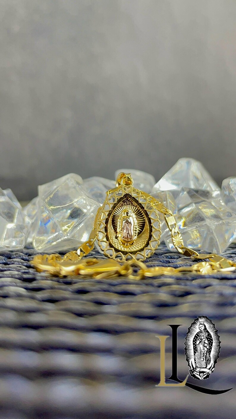 Puede incluir: Un collar con colgante dorado que representa a la Virgen Mar&iacute;a. El colgante tiene un dise&ntilde;o calado y ornamentado y cuelga de una cadena a juego. El collar se exhibe sobre una superficie azul texturizada con detalles transparentes en forma de cristal.
