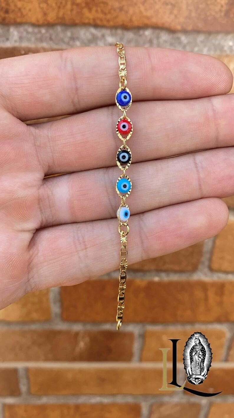 May include: Gold chain bracelet with five evil eye charms. The charms are blue, red, black, light blue, and white.