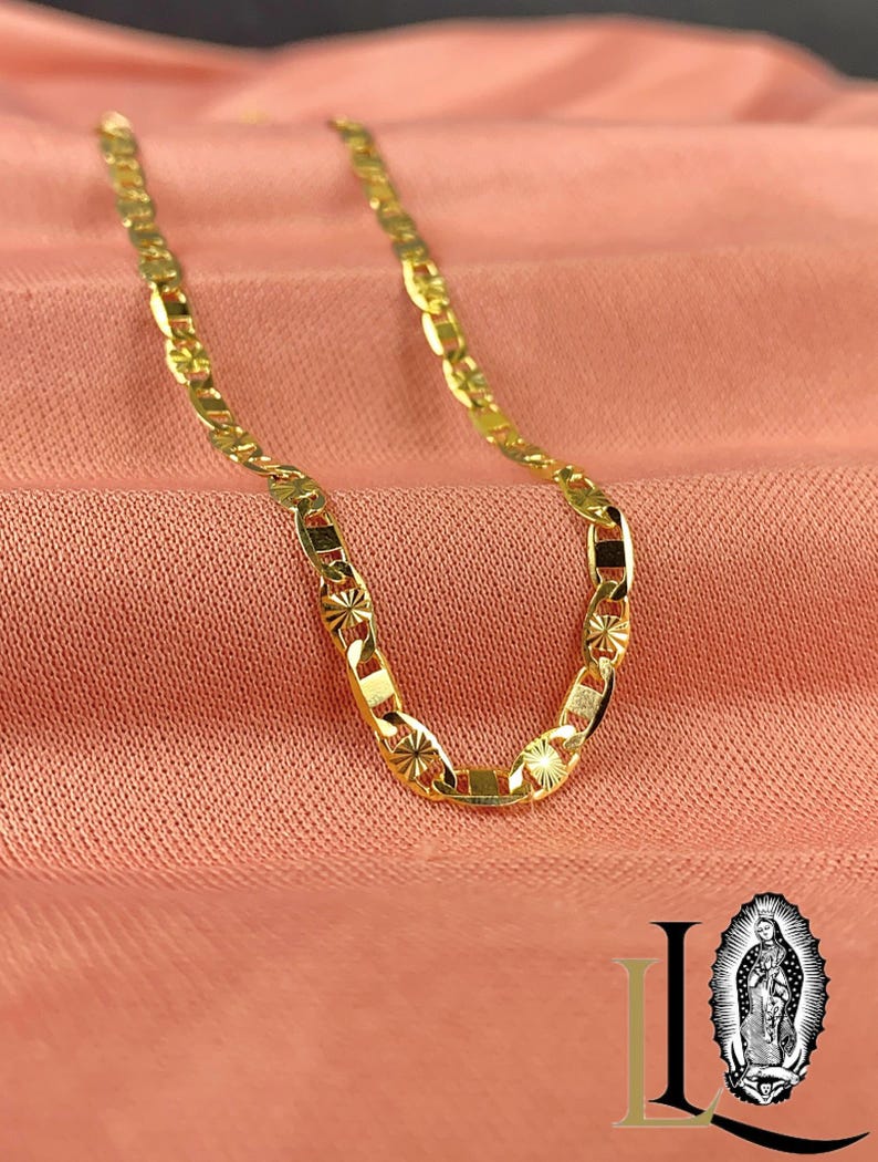 May include: A gold-colored chain necklace with a unique link design, resting on a peach-colored fabric. The chain features starburst and rectangular details. The image also includes a logo with the image of the Virgin Mary.
