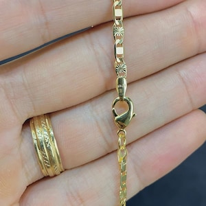 May include: A gold-colored necklace with a lobster clasp, shown against a dark background. The chain has a mix of rectangular and decorative links. A gold ring is visible on a finger. The necklace is likely made of gold or gold-plated material.