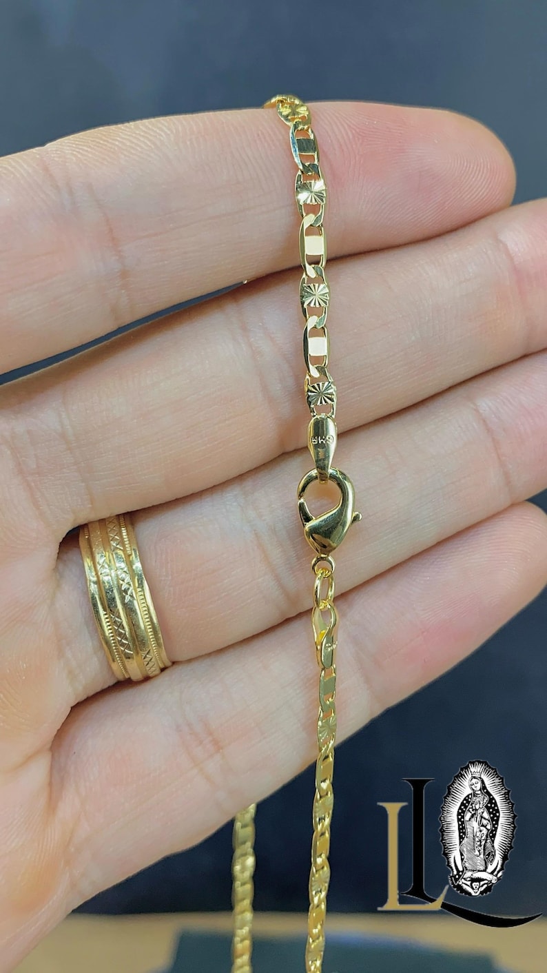 May include: A gold-colored necklace with a lobster clasp, shown against a dark background. The chain has a mix of rectangular and decorative links. A gold ring is visible on a finger. The necklace is likely made of gold or gold-plated material.