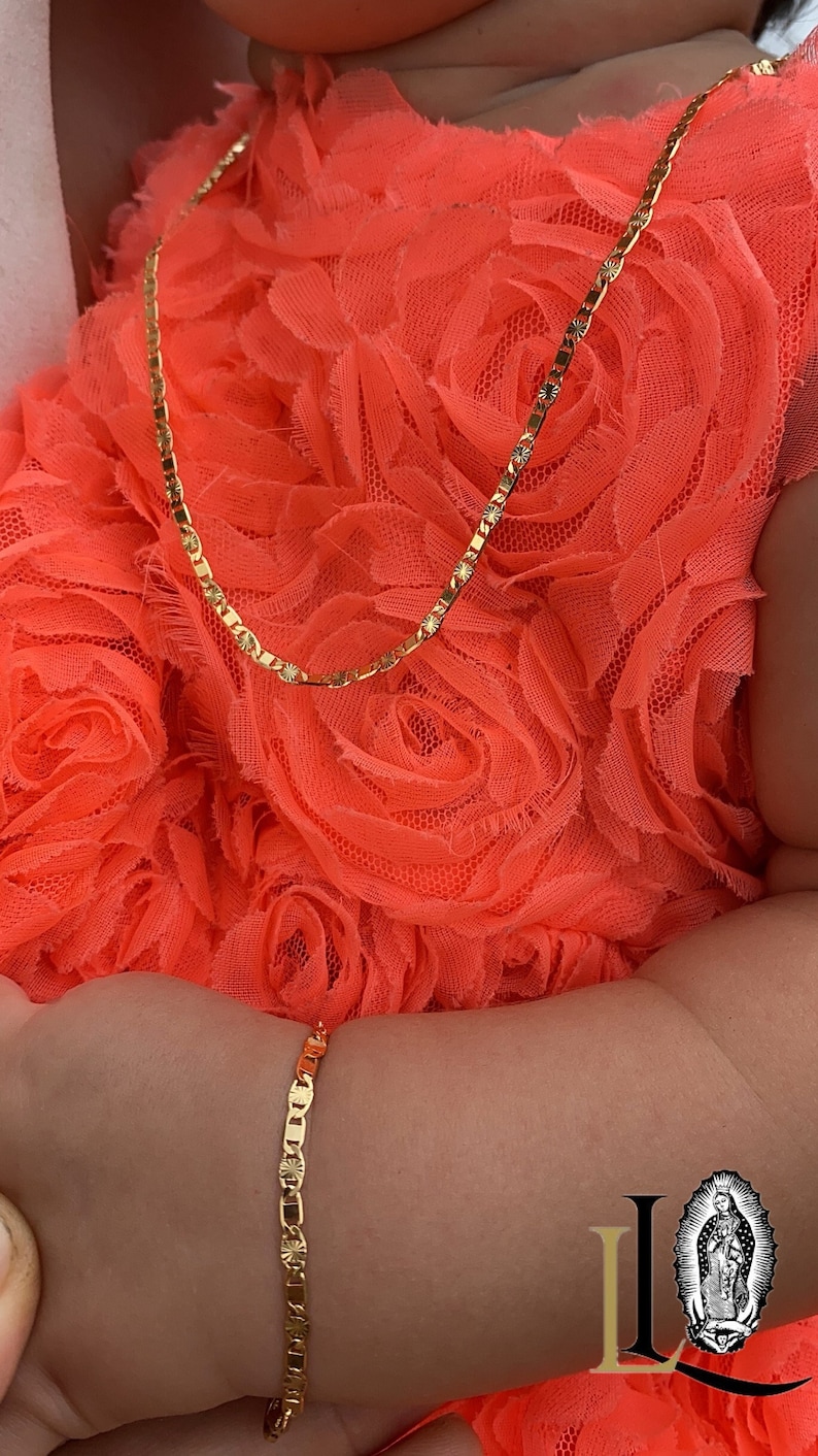 Puede incluir: Un conjunto de collar y pulsera de color dorado. El collar y la pulsera tienen un dise&ntilde;o delicado de eslabones planos con detalles de estrellas. Las joyas se exhiben sobre un fondo de tela naranja brillante y con volantes.