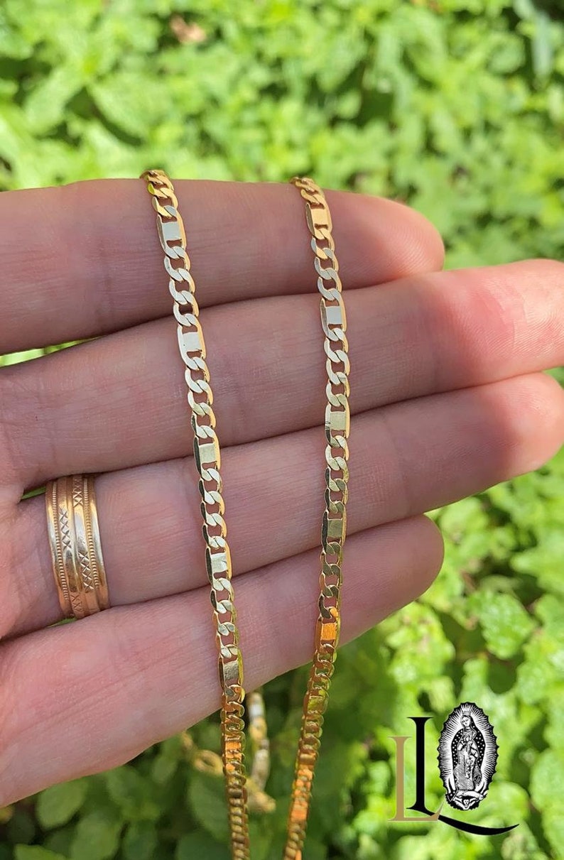 May include: Two gold chains with a curb link pattern. The chains are laid out side by side on a hand.