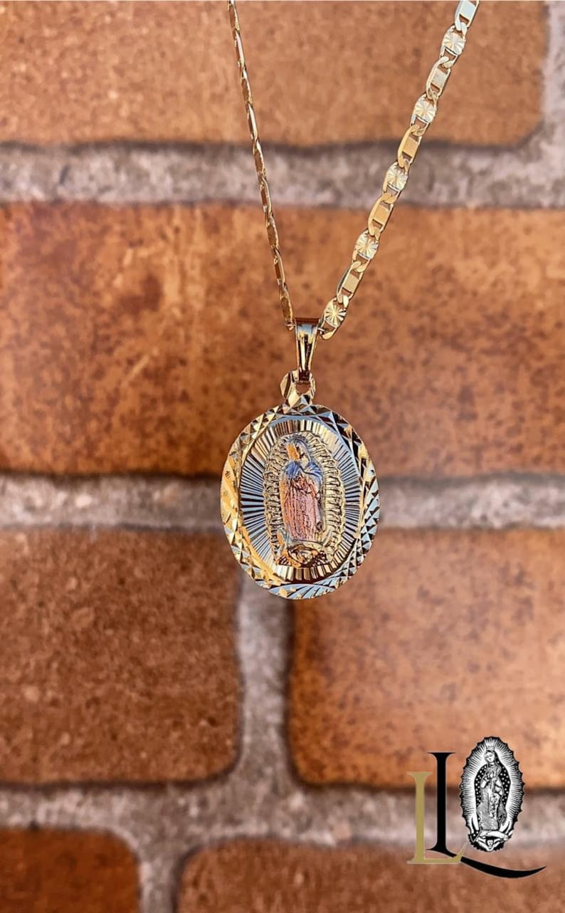 May include: A gold chain necklace with a two-tone oval pendant featuring an image of the Virgin Mary. The pendant has a diamond-shaped pattern.