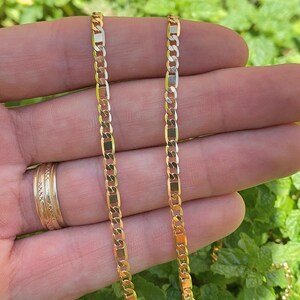 May include: A gold chain necklace with a curb link style. The chain is shown on a hand against a green background.