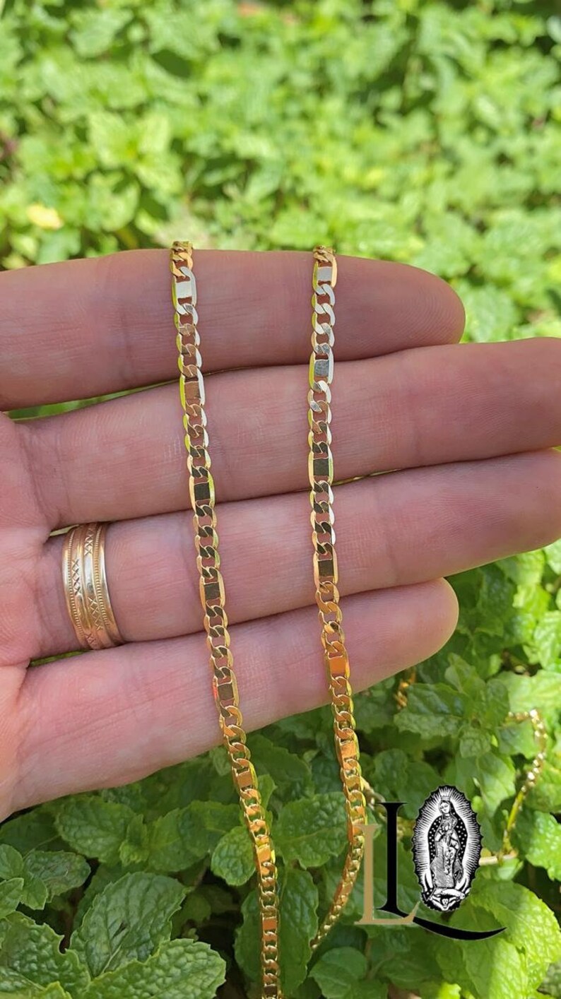 May include: A gold chain necklace with a curb link style. The chain is shown on a hand against a green background.