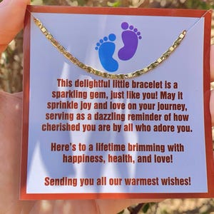 May include: A gold chain bracelet with a small charm in the shape of two footprints. The bracelet is on a card with the text "This delightful little bracelet is a sparkling gem, just like you! May it sprinkle joy and love on your journey, serving as a dazzling reminder of how cherished you are by all who adore you. Here's to a lifetime brimming with happiness, health, and love! Sending you all our warmest wishes!"