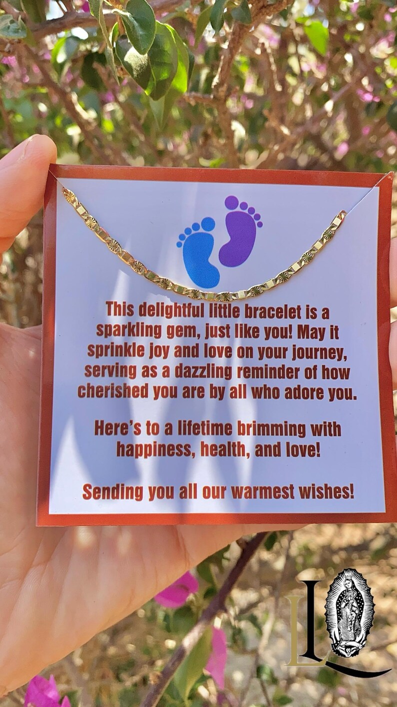 May include: A gold chain bracelet with a small charm in the shape of two footprints. The bracelet is on a card with the text "This delightful little bracelet is a sparkling gem, just like you! May it sprinkle joy and love on your journey, serving as a dazzling reminder of how cherished you are by all who adore you. Here's to a lifetime brimming with happiness, health, and love! Sending you all our warmest wishes!"