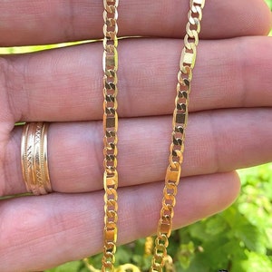 May include: Two gold chains, one on top of the other, are shown in a close-up image. The chains are both made of a flat, oval-shaped link. The chains are lying on a person's hand.