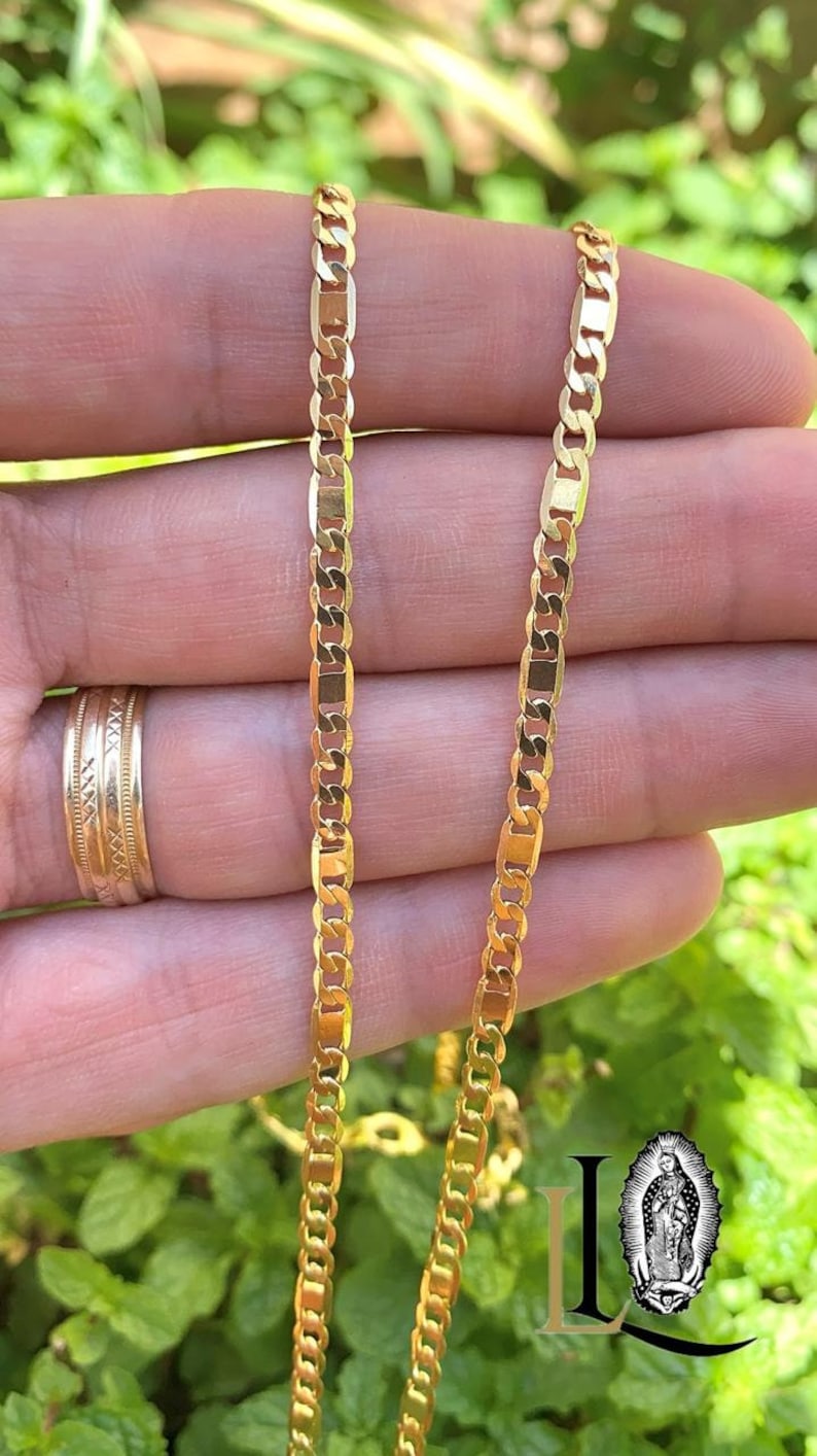 May include: Two gold chains, one on top of the other, are shown in a close-up image. The chains are both made of a flat, oval-shaped link. The chains are lying on a person's hand.