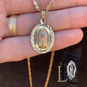May include: A gold chain necklace with a pendant featuring a depiction of the Virgin Mary. The pendant is oval-shaped and has a diamond-cut design. The Virgin Mary is depicted in a standing pose with her hands clasped in prayer. The pendant is made of two-toned gold, with the Virgin Mary's image in rose gold and the rest of the pendant in yellow gold.