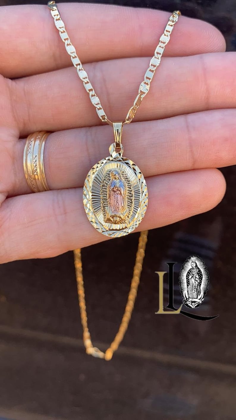 May include: A gold chain necklace with a pendant featuring a depiction of the Virgin Mary. The pendant is oval-shaped and has a diamond-cut design. The Virgin Mary is depicted in a standing pose with her hands clasped in prayer. The pendant is made of two-toned gold, with the Virgin Mary's image in rose gold and the rest of the pendant in yellow gold.