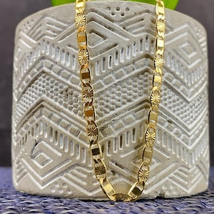 May include: A gold-colored chain necklace with a repeating pattern of rectangular and star-shaped links. The necklace is displayed on a textured gray pot with a green plant. The background is a blue surface.
