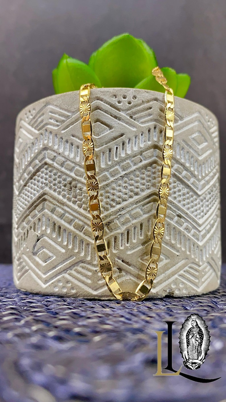 May include: A gold-colored chain necklace with a repeating pattern of rectangular and star-shaped links. The necklace is displayed on a textured gray pot with a green plant. The background is a blue surface.