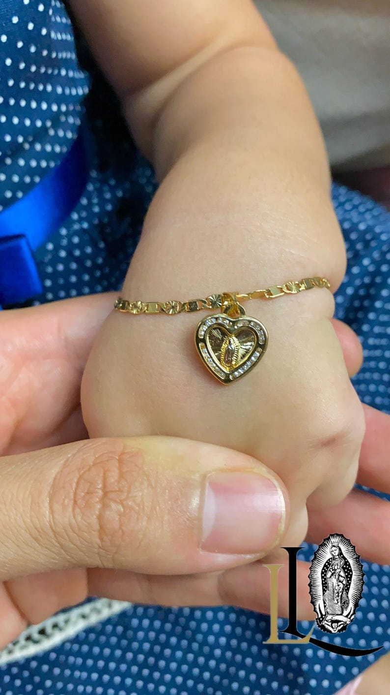 May include: A gold bracelet with a heart-shaped pendant. The pendant features a religious figure and is set with small, clear stones. The bracelet is on a child's wrist, with a blue polka-dot background.