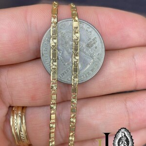 May include: Close-up of a gold-colored chain necklace with a rectangular and starburst design. The chain is held next to a quarter coin for size comparison. The chain is a bright, shiny gold color.