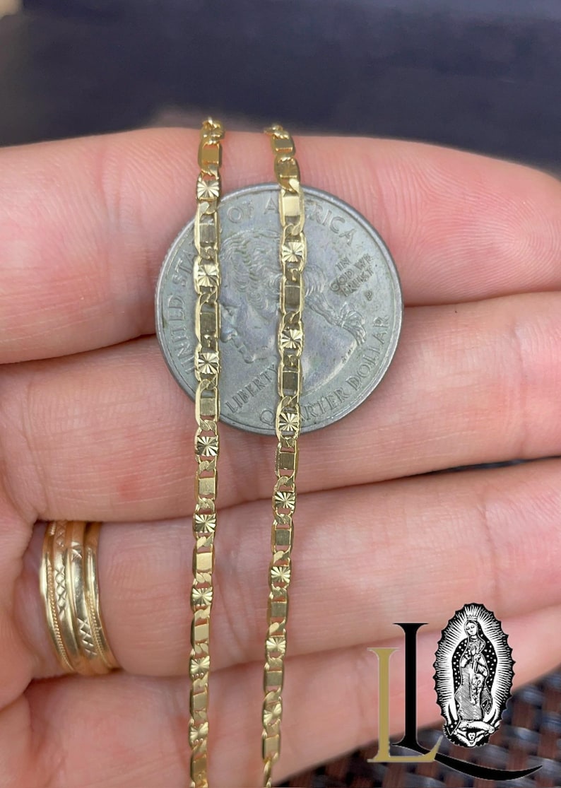 May include: Close-up of a gold-colored chain necklace with a rectangular and starburst design. The chain is held next to a quarter coin for size comparison. The chain is a bright, shiny gold color.