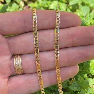 May include: Two gold chains, one slightly longer than the other, are shown in a close-up image. The chains are made of a flat, curb link design. The chains are held in a person's hand against a background of green foliage.