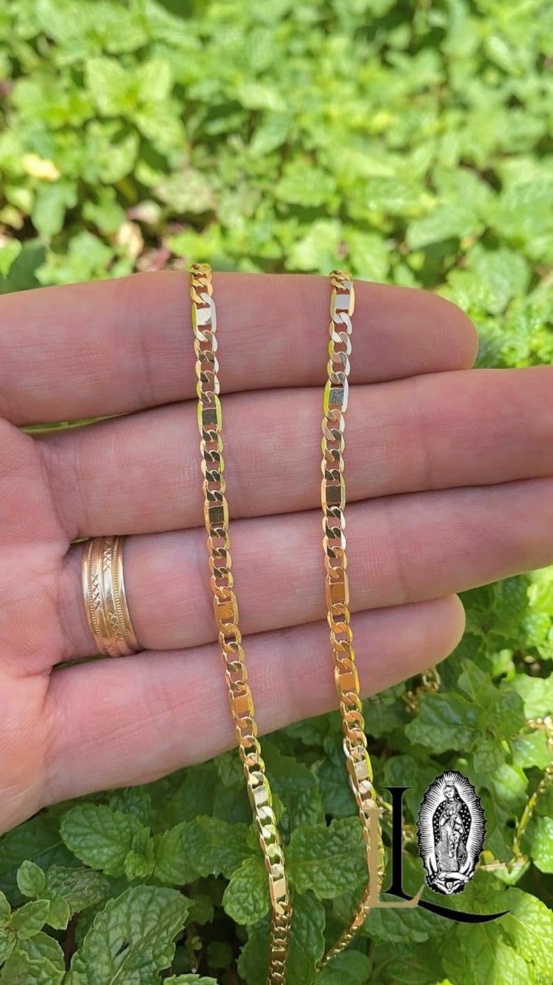 May include: Two gold chains, one slightly longer than the other, are shown in a close-up image. The chains are made of a flat, curb link design. The chains are held in a person's hand against a background of green foliage.
