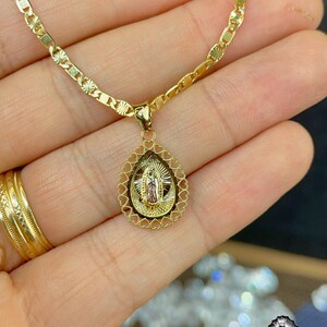 Puede incluir: Un collar dorado con un colgante que representa una figura religiosa. El colgante tiene forma de l&aacute;grima, con un borde en forma de coraz&oacute;n. La cadena es de dise&ntilde;o delicado de eslabones planos. El collar se sostiene en una mano.