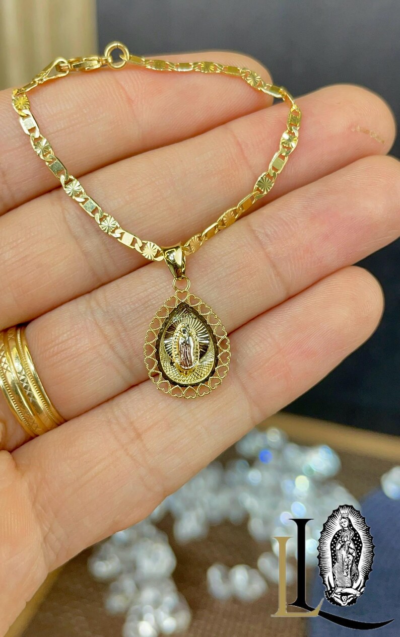 Puede incluir: Un collar dorado con un colgante que representa una figura religiosa. El colgante tiene forma de l&aacute;grima, con un borde en forma de coraz&oacute;n. La cadena es de dise&ntilde;o delicado de eslabones planos. El collar se sostiene en una mano.