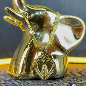 May include: A gold necklace with a heart-shaped pendant depicting a religious figure, adorned with clear stones. The necklace is displayed on a gold elephant ornament. The chain is a thick curb style.