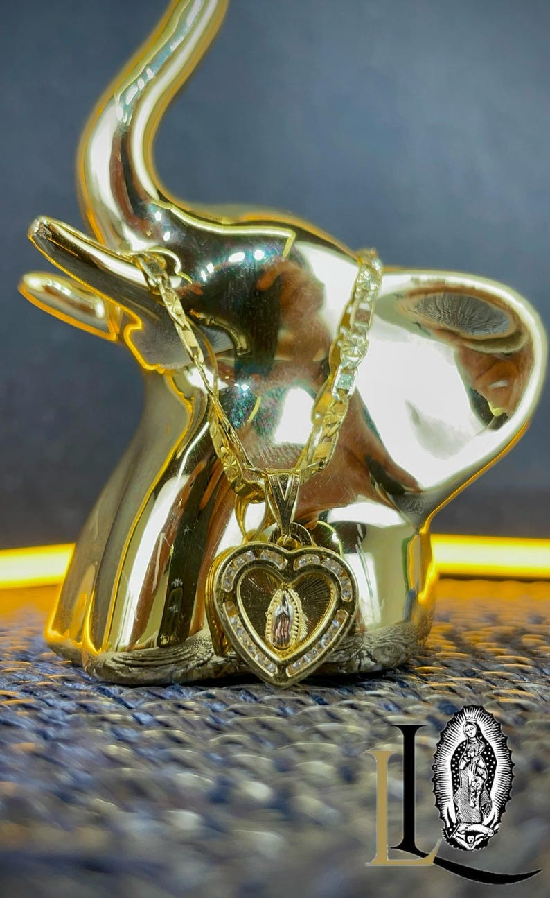 May include: A gold necklace with a heart-shaped pendant depicting a religious figure, adorned with clear stones. The necklace is displayed on a gold elephant ornament. The chain is a thick curb style.