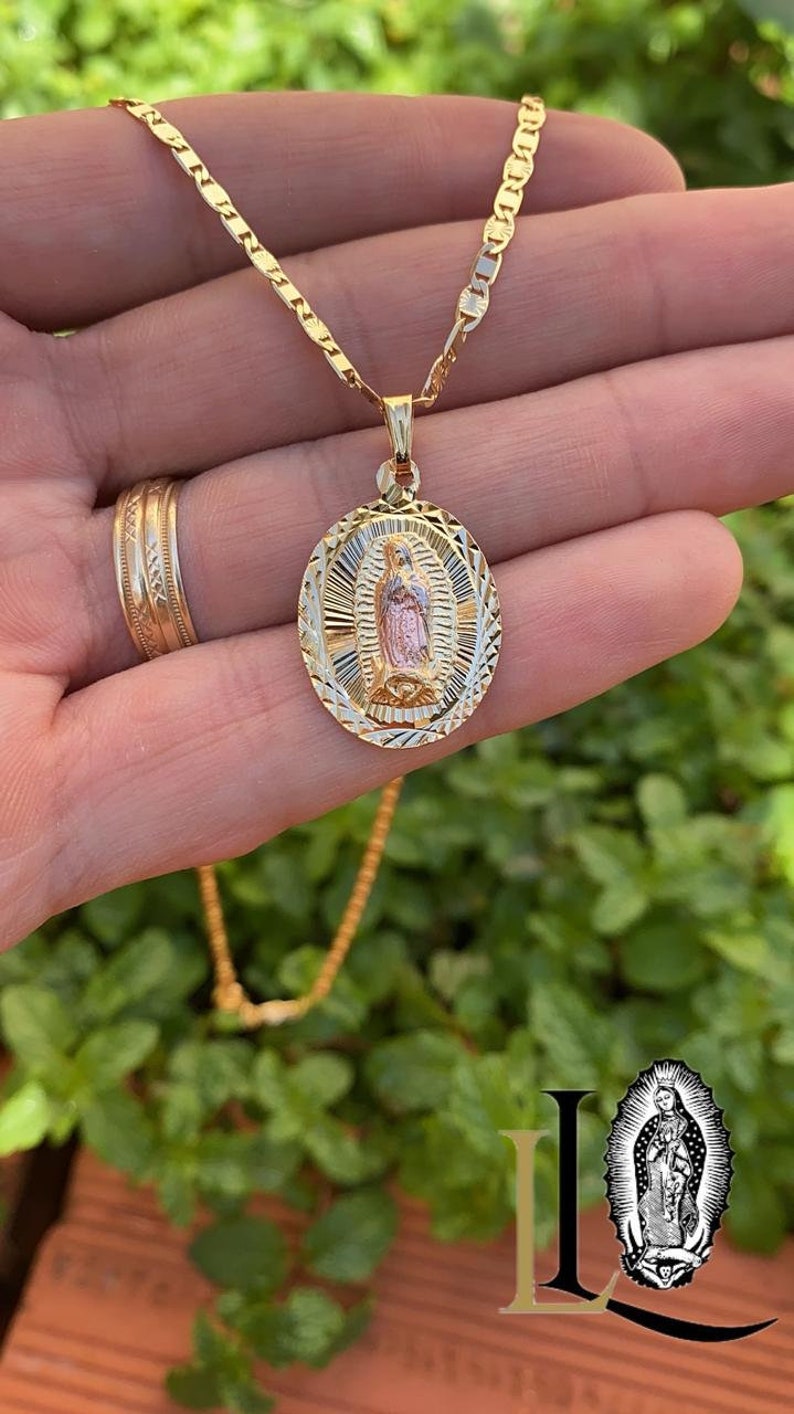 May include: A gold chain necklace with a two-tone oval pendant featuring a depiction of the Virgin Mary. The pendant has a detailed, textured design.
