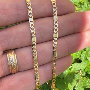 May include: Two gold chains, one thicker than the other, are shown in a hand. The chains are both made of a flat, rectangular link design.