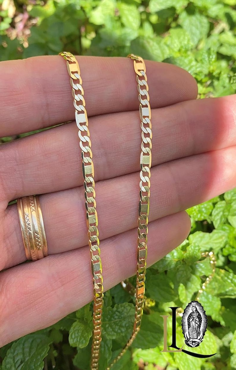 May include: Two gold chains, one thicker than the other, are shown in a hand. The chains are both made of a flat, rectangular link design.