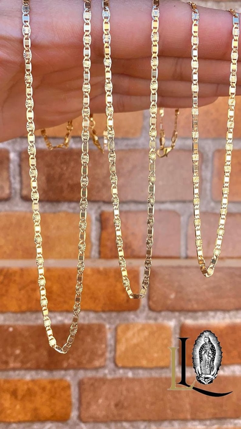 May include: Four gold chains with a delicate, patterned design. The chains are laid out on a hand and a brick wall background.