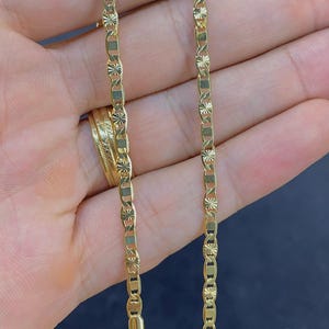 May include: A gold-colored chain necklace with a unique link design. The chain is held in a hand, showcasing its intricate pattern and the clasp. The necklace appears to be made of a shiny metal, reflecting light.