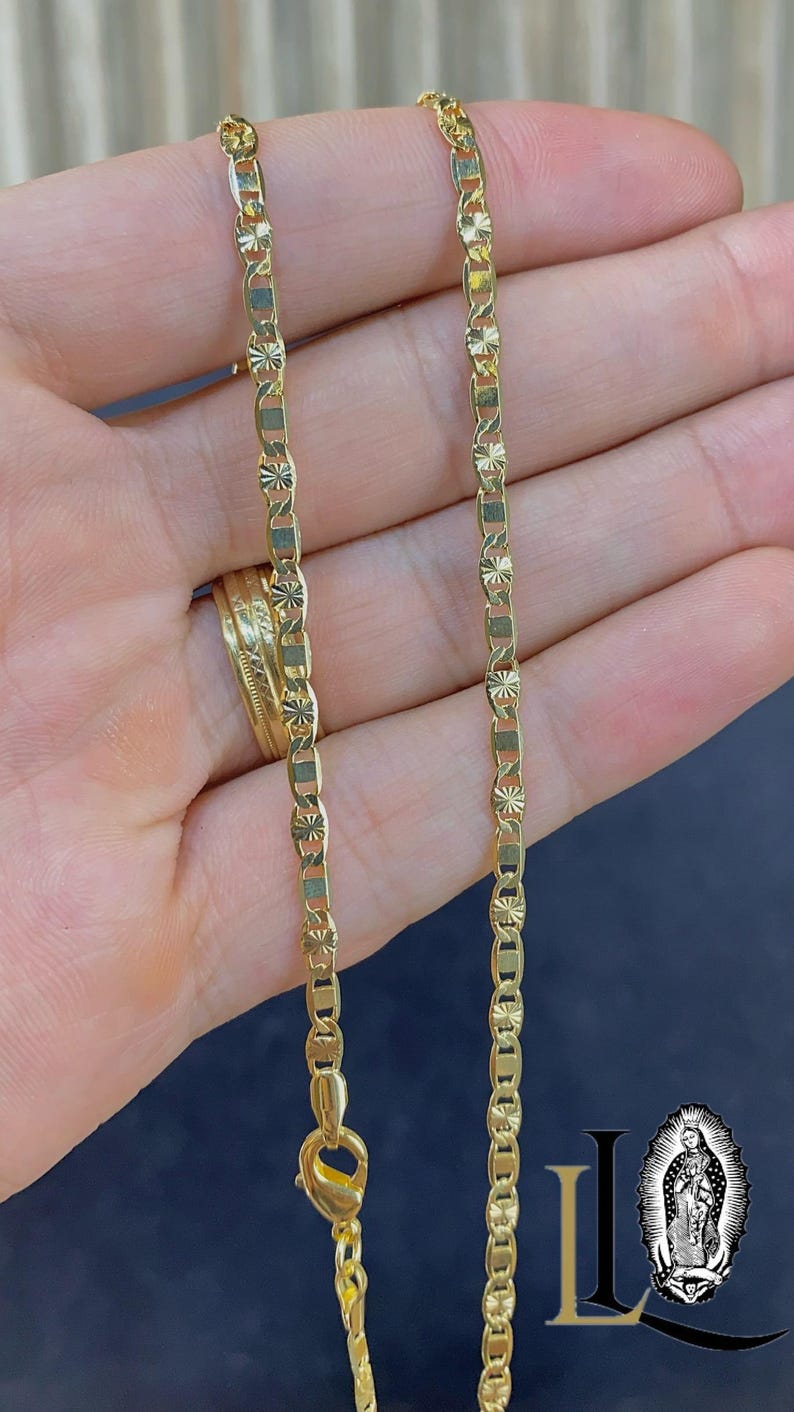 May include: A gold-colored chain necklace with a unique link design. The chain is held in a hand, showcasing its intricate pattern and the clasp. The necklace appears to be made of a shiny metal, reflecting light.