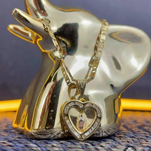 May include: A gold elephant ornament with a gold heart pendant necklace. The pendant has a religious image and is set with clear stones. The necklace is draped over the elephant's trunk. The background is a textured blue surface.