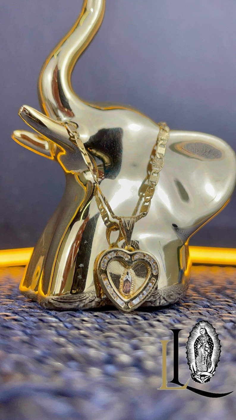 May include: A gold elephant ornament with a gold heart pendant necklace. The pendant has a religious image and is set with clear stones. The necklace is draped over the elephant's trunk. The background is a textured blue surface.