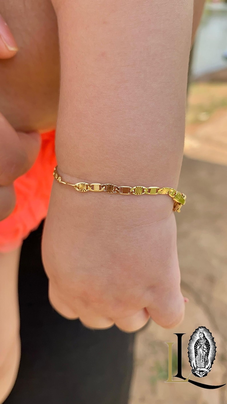 May include: A gold chain bracelet with a delicate design. The bracelet is on a child's wrist.