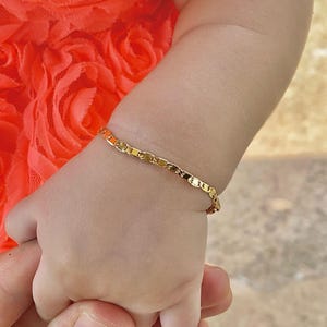 May include: A close-up of a baby's arm wearing a gold chain bracelet. The baby is wearing a bright orange floral dress.