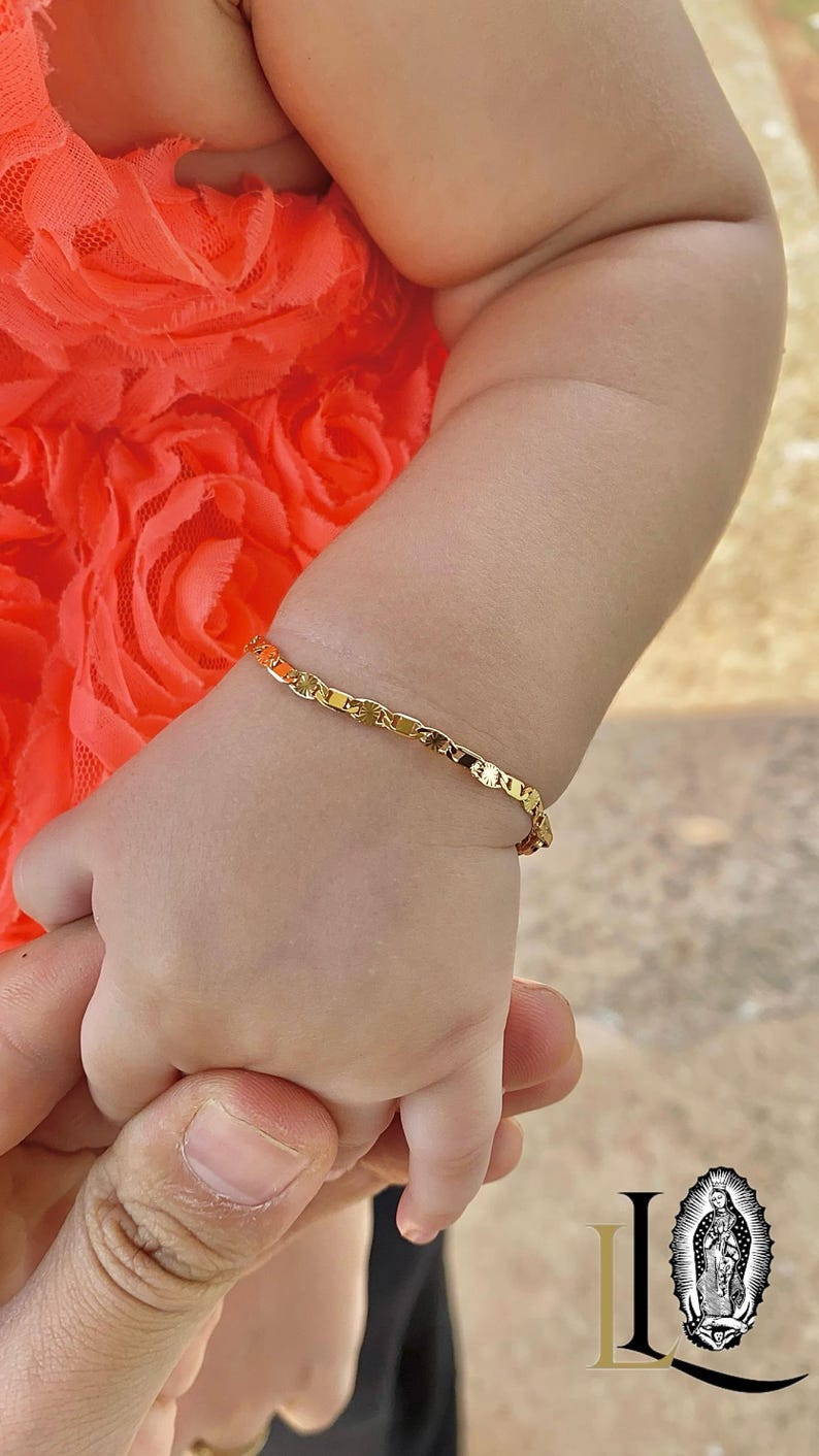 May include: A close-up of a baby's arm wearing a gold chain bracelet. The baby is wearing a bright orange floral dress.